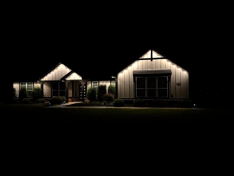 White board-and-batten farmhouse with warm white permanent roofline lighting on every gable